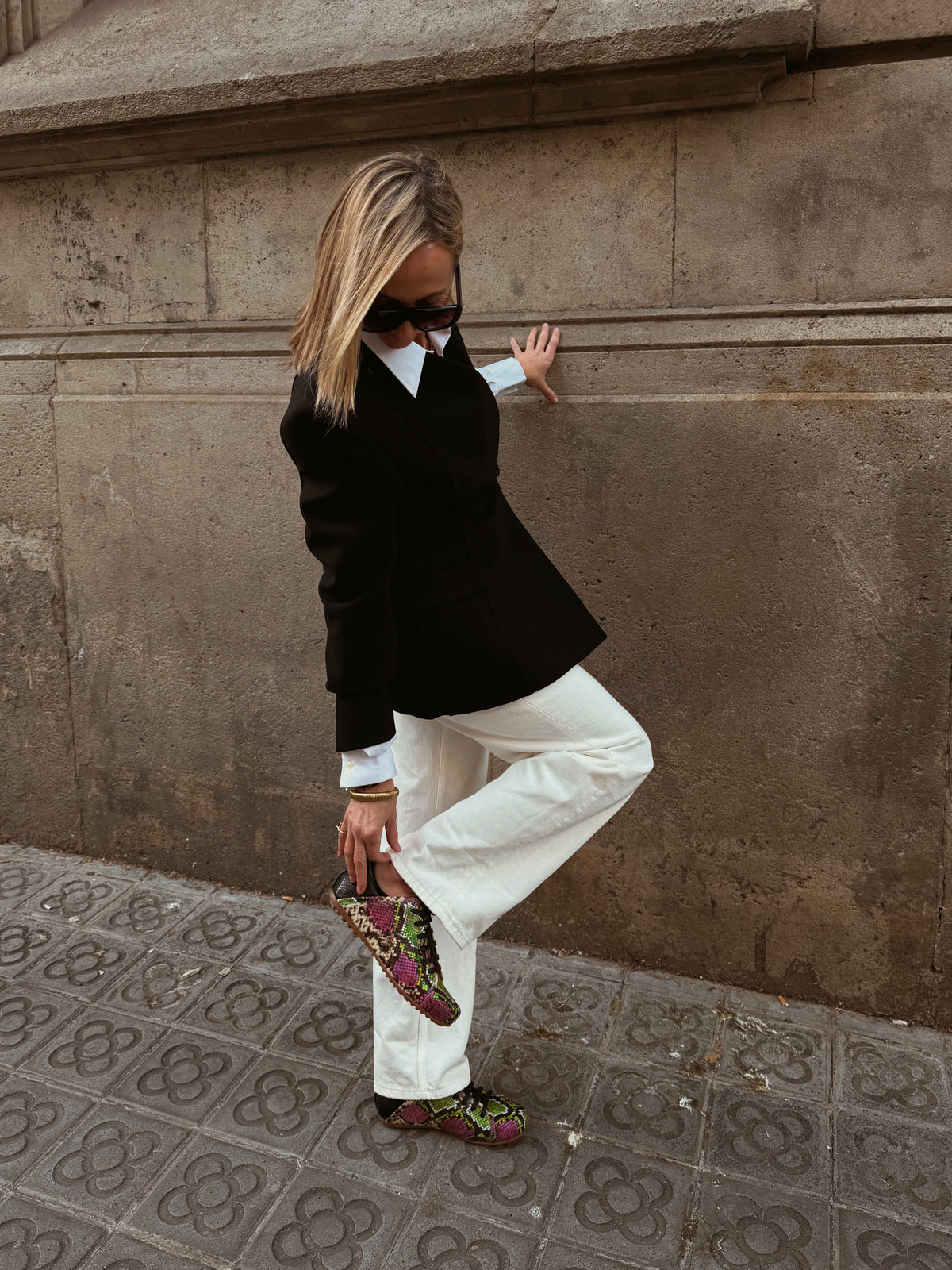 A woman posing outdoors against a textured stone wall, wearing a black blazer, white collared shirt, white wide-leg trousers, and distinctive green and pink patterned loafers.